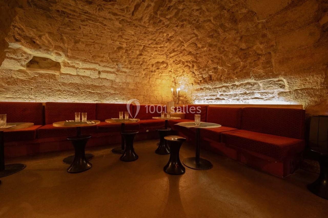 Banquettes rouges et tables rondes dans une salle voûtée en pierre éclairée par une lumière tamisée.