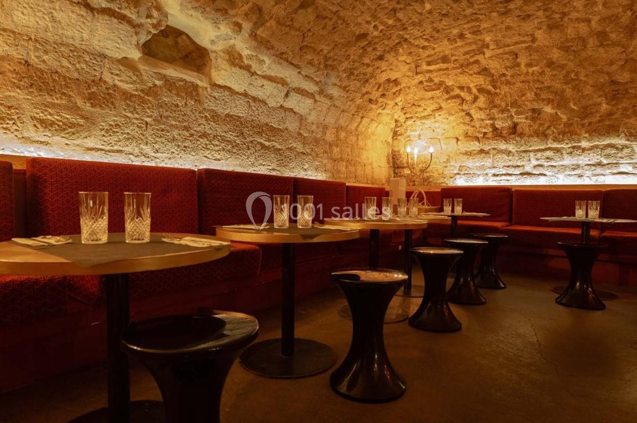 Salle voûtée en pierre avec banquettes rouges, tables en bois, tabourets noirs et verres disposés.