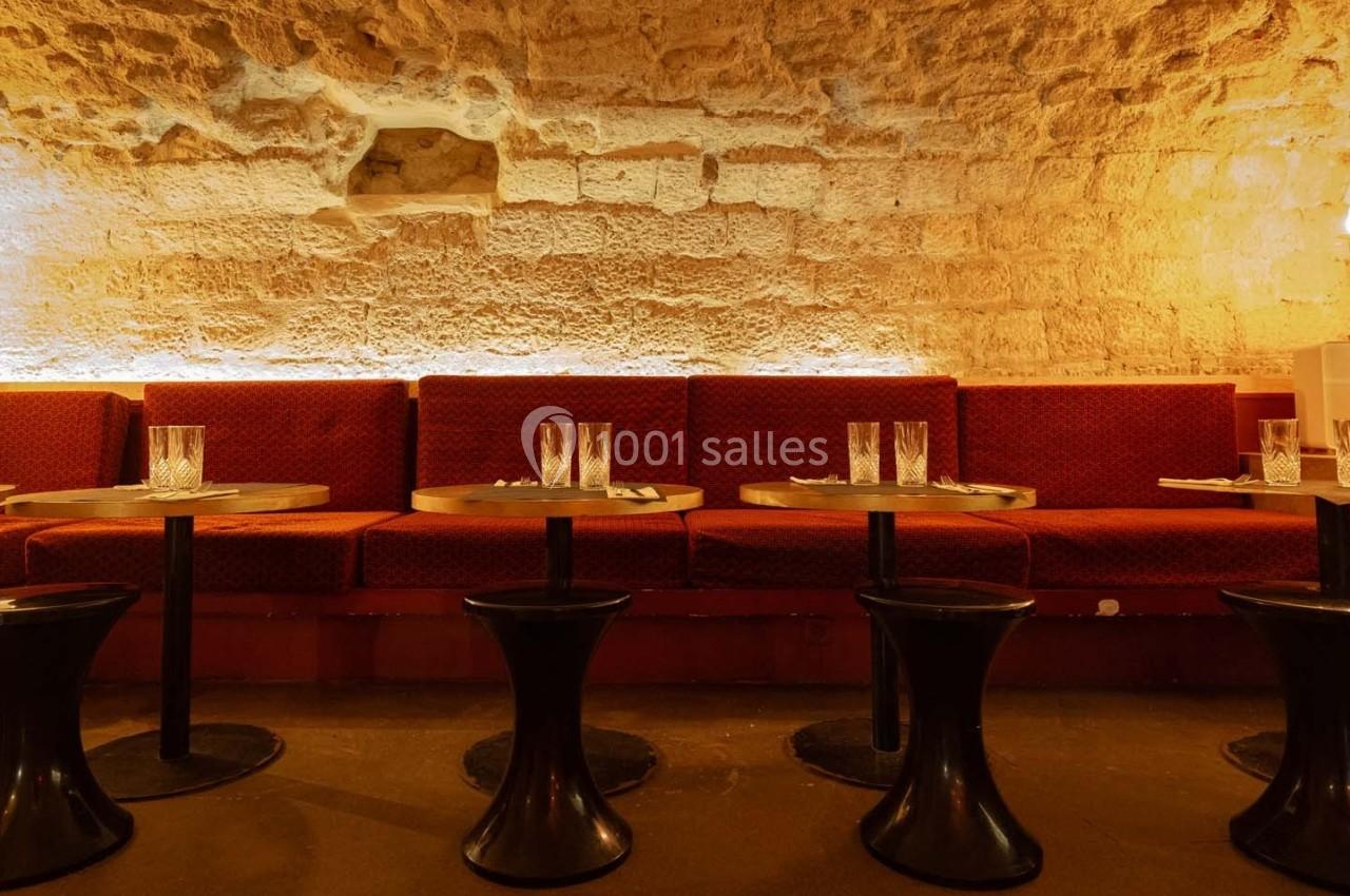 Banquettes rouges et tables rondes avec verres, dans une salle voûtée en pierre éclairée par une lumière chaude.