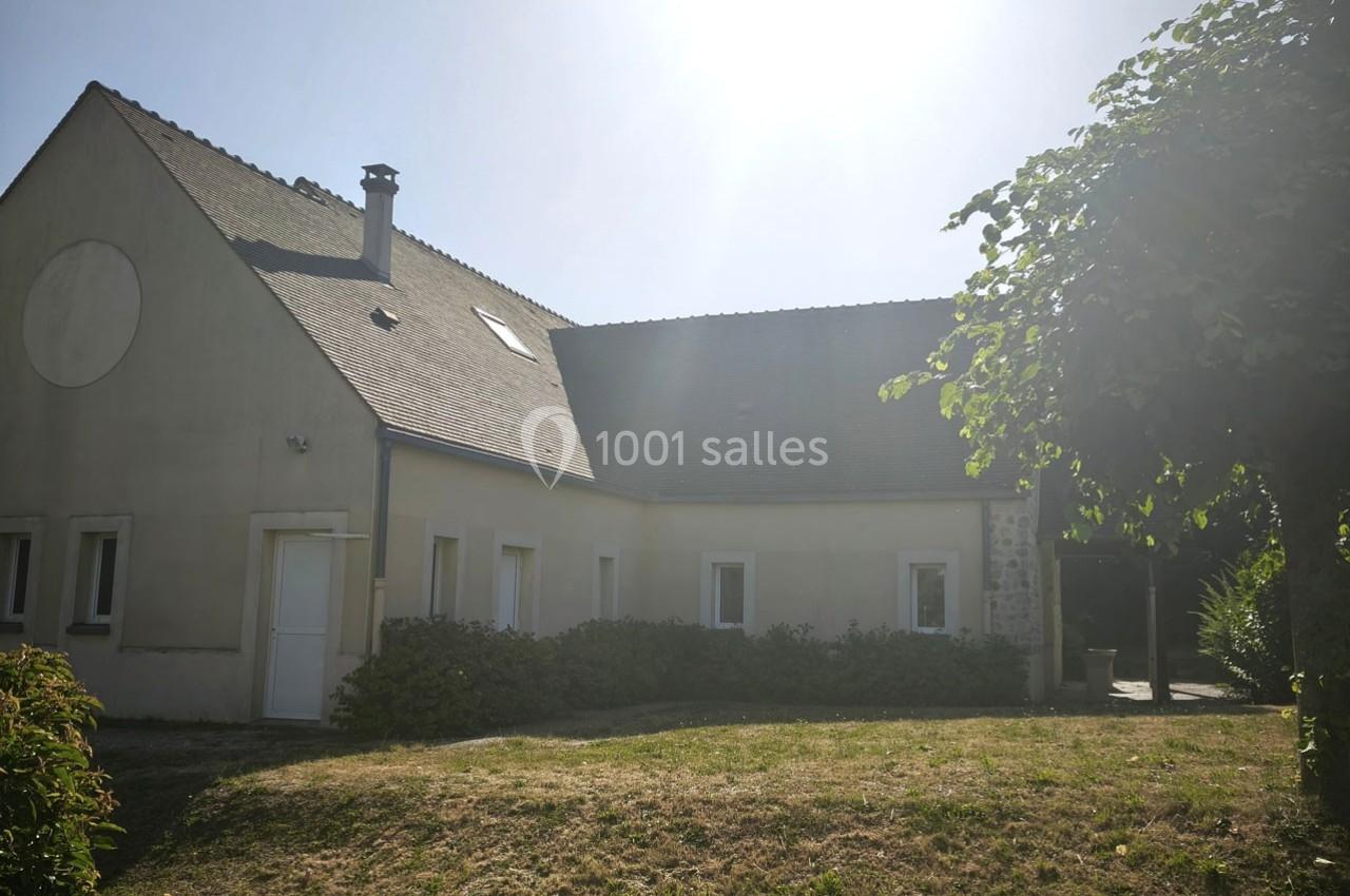 Maison ensoleillée avec toit en pente, façade beige, entourée de verdure et d'arbustes sur une pelouse.