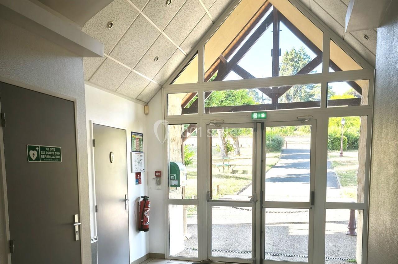 Entrée vitrée d'un bâtiment avec porte double, panneau de sortie de secours et vue sur un chemin extérieur arboré.
