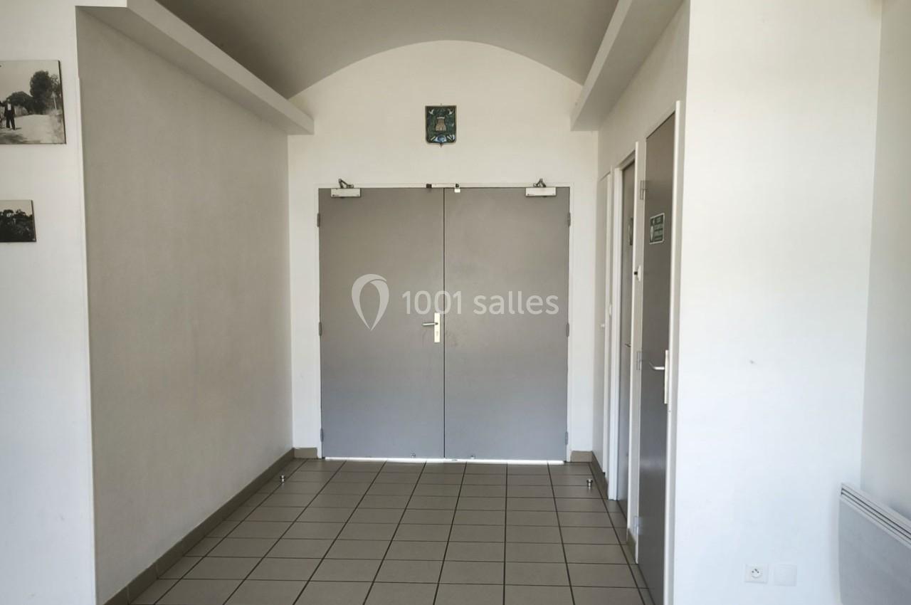 Porte double grise dans un couloir lumineux avec murs blancs, sol carrelé et panneaux indiquant des toilettes.