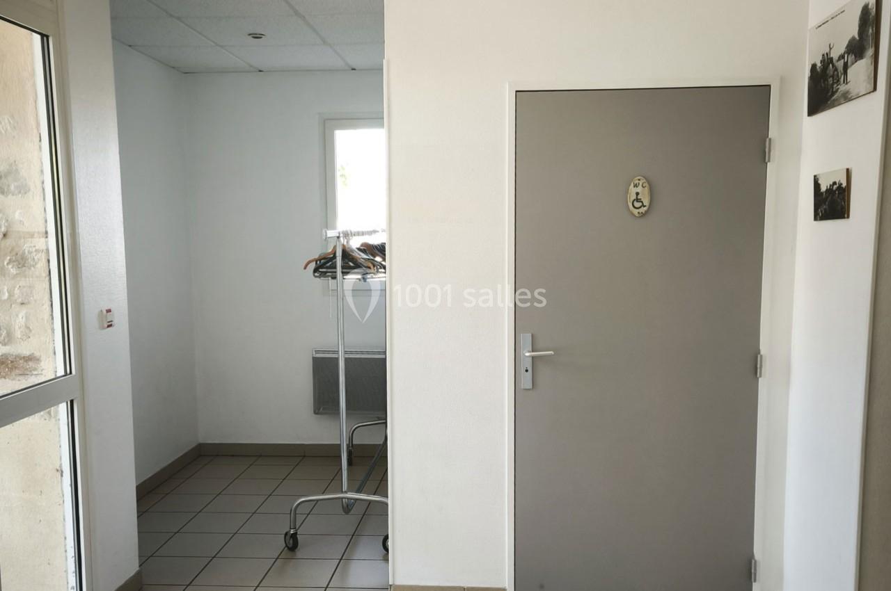 Porte grise avec pictogramme, portant à vêtements sur roulettes et fenêtre dans un couloir carrelé.