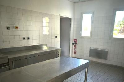 Cuisine professionnelle vide avec plans de travail en inox, murs carrelés blancs, fenêtres et extincteur visible.