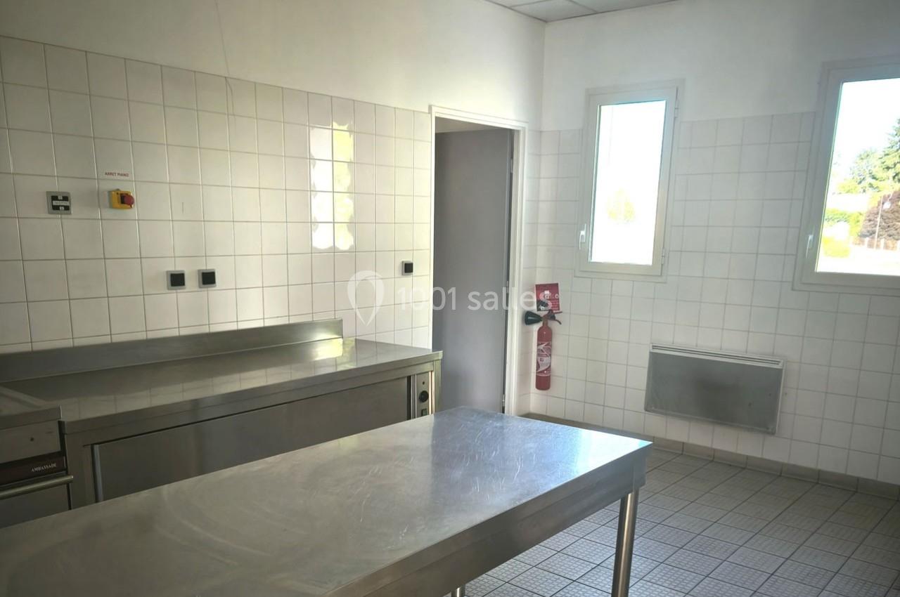 Cuisine professionnelle vide avec plans de travail en inox, murs carrelés blancs, fenêtres et extincteur visible.