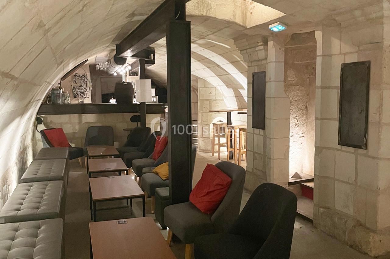 Salle voûtée en pierre avec canapés, fauteuils, tables basses et un bar en arrière-plan.