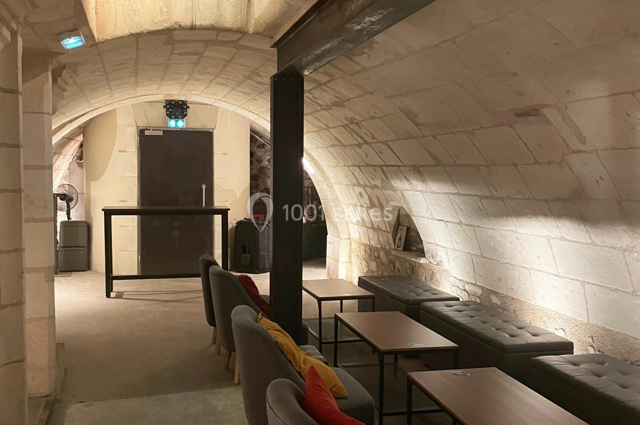 Salle voûtée en pierre avec des tables, des fauteuils gris et une lumière tamisée.