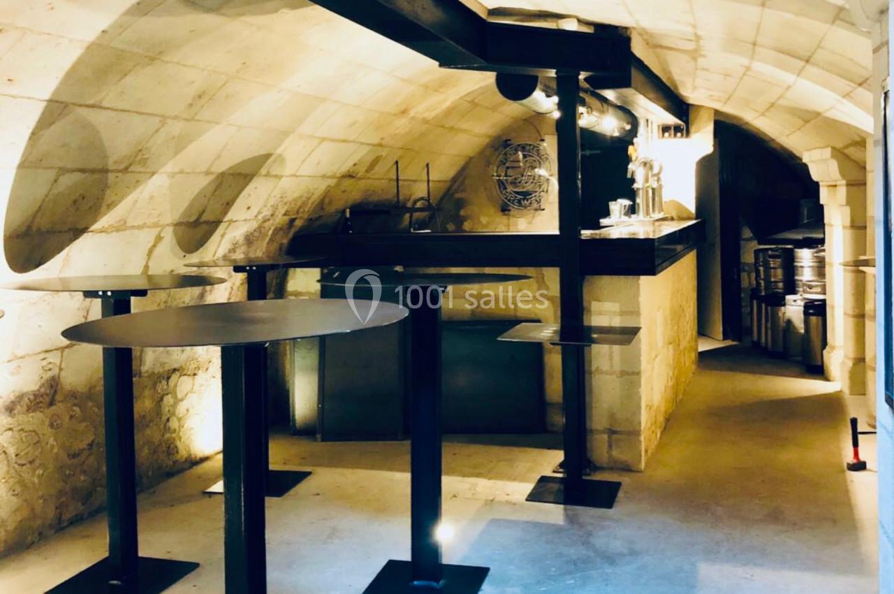 Salle voûtée en pierre avec tables hautes et comptoir, éclairage chaleureux et ambiance sobre.