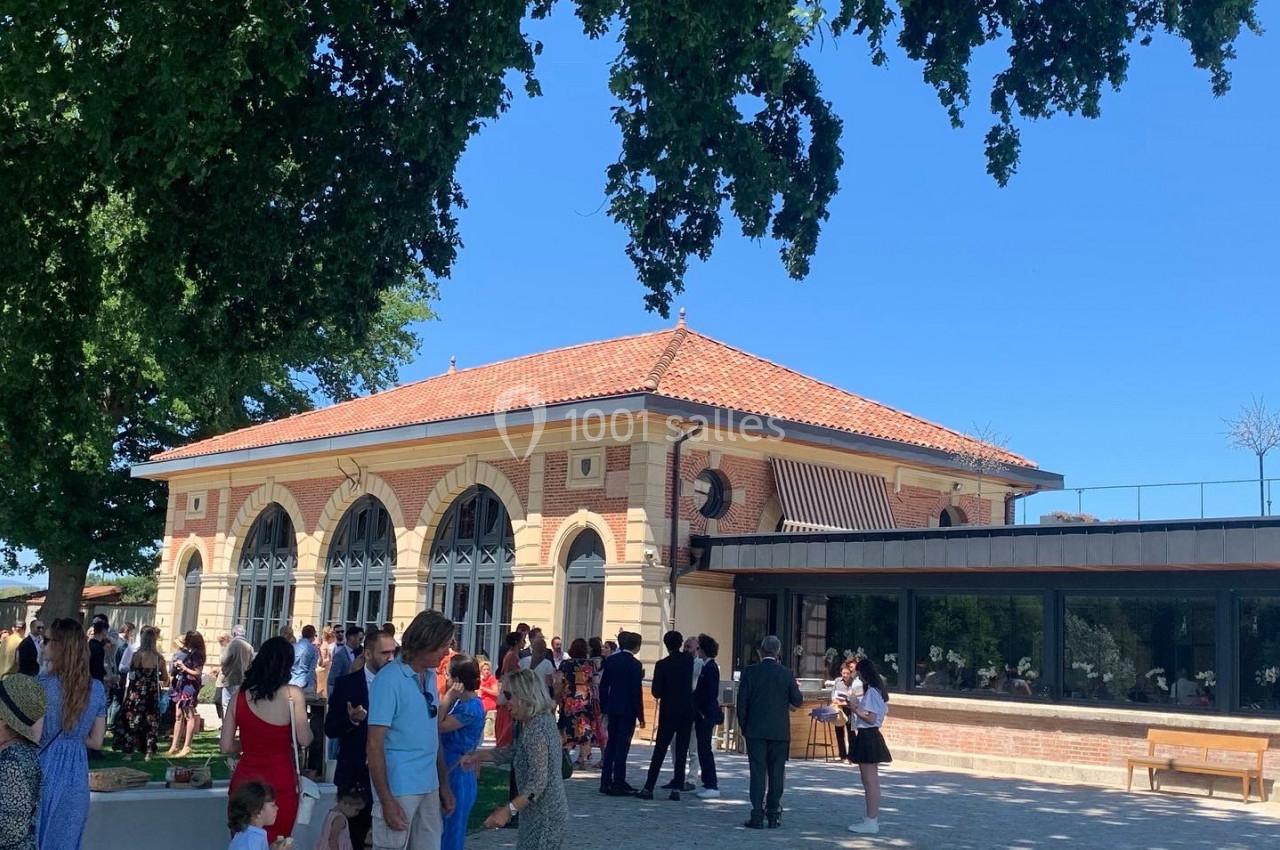 Groupe de personnes rassemblées devant un bâtiment en briques avec de grandes arches, sous un ciel bleu et des arbres.