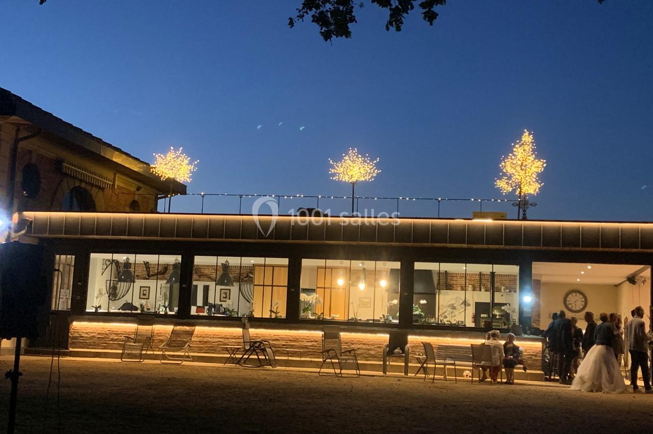 Façade d'un bâtiment éclairé avec des guirlandes lumineuses et des arbres décorés, en soirée, avec des personnes rassemblées.