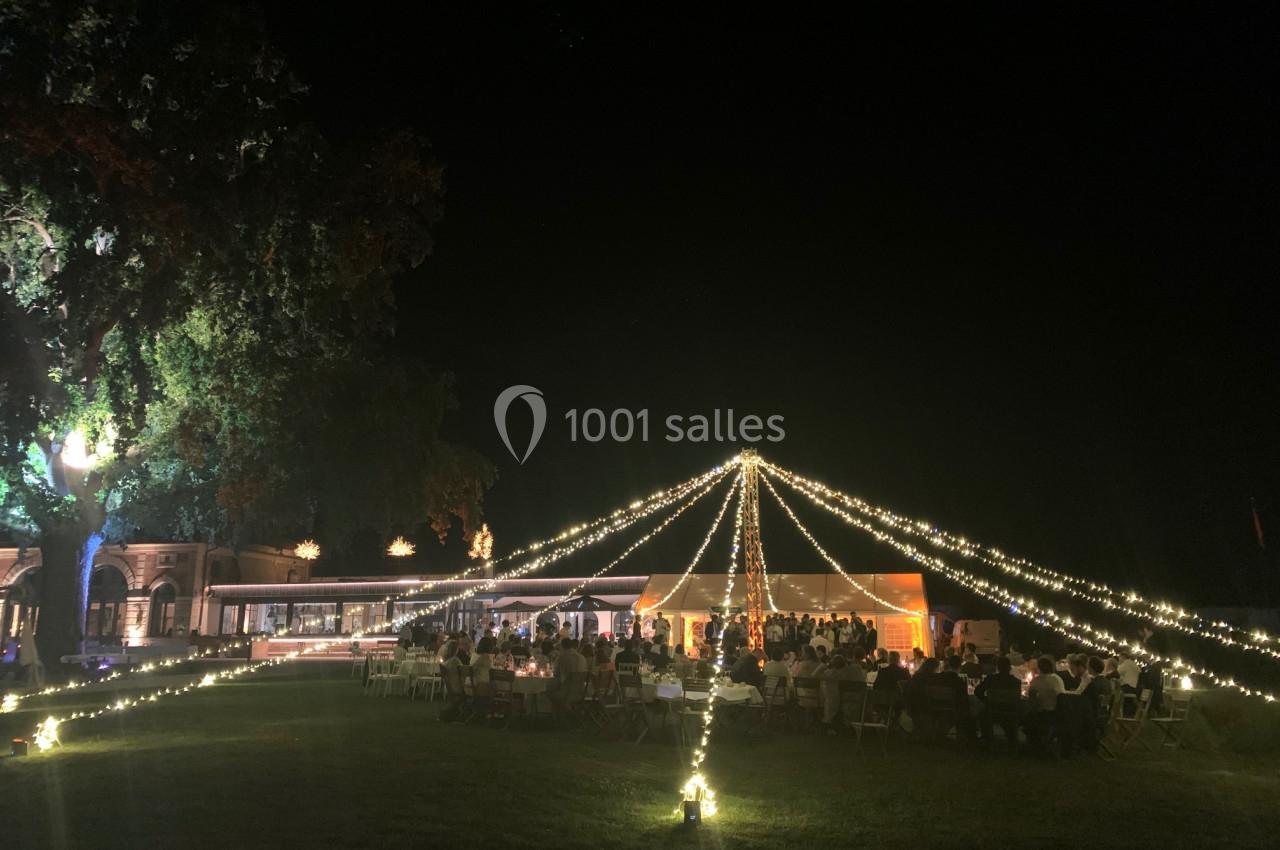 Tente éclairée par des guirlandes lumineuses dans un jardin, entourée de convives en soirée.