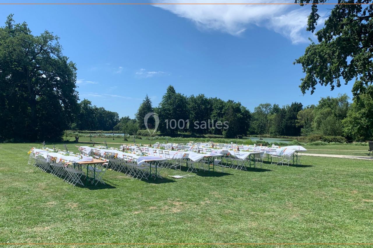 Tables et chaises blanches disposées en extérieur sur une pelouse entourée d'arbres sous un ciel partiellement dégagé.