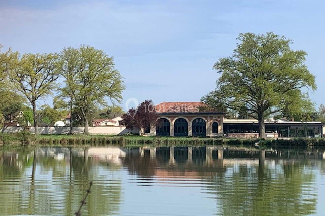 Bâtiment en pierre avec arches, situé au bord d'un étang entouré d'arbres et de végétation.