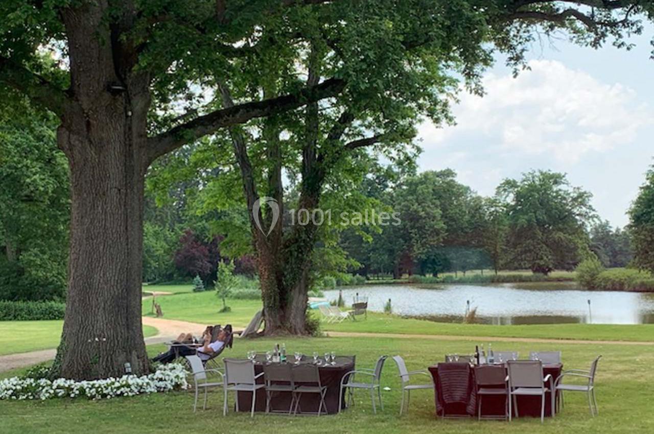 Tables et chaises disposées sur une pelouse ombragée près d'un étang, entourées d'arbres et de verdure.