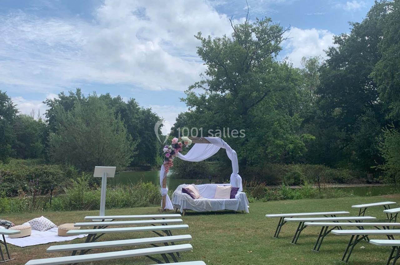 Cérémonie en plein air avec arche fleurie blanche et violette, bancs alignés sur une pelouse près d'un étang.