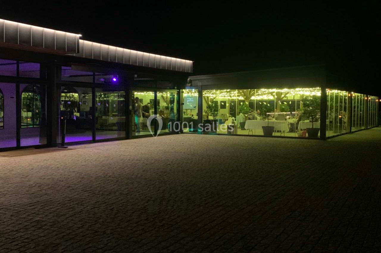 Salle de réception vitrée éclairée de nuit, avec des tables dressées visibles à l'intérieur.