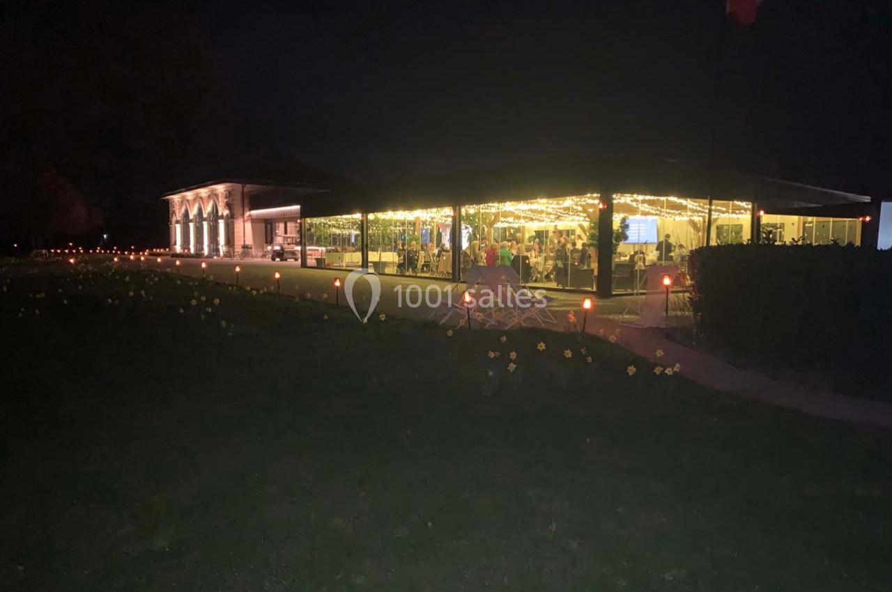 Bâtiment éclairé par des guirlandes lumineuses la nuit, entouré d'un jardin sombre avec des fleurs et des bougies.