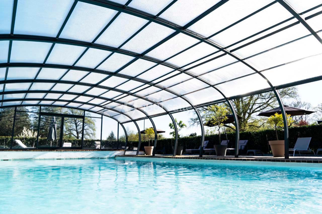 Piscine couverte avec une structure en verre incurvée, entourée de chaises longues et de végétation.