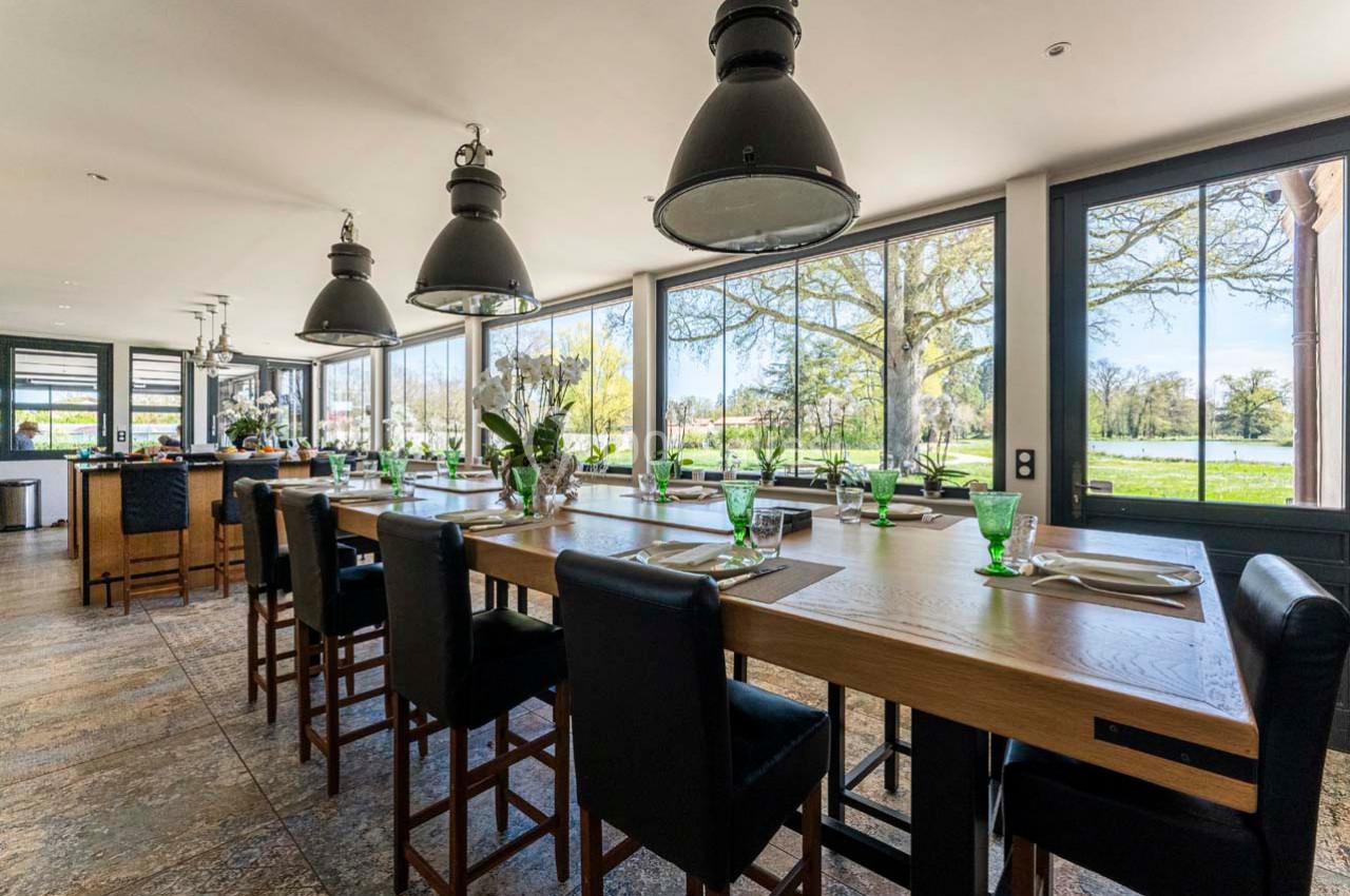 Salle lumineuse avec une grande table en bois, chaises hautes, vaisselle verte et vue sur un paysage verdoyant.