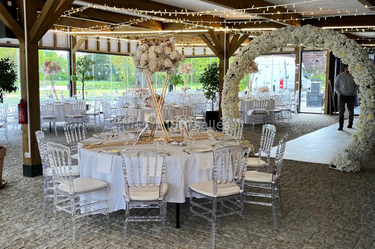 Salle de réception décorée avec des tables rondes, des chaises transparentes et une arche florale sous un plafond lumineux.