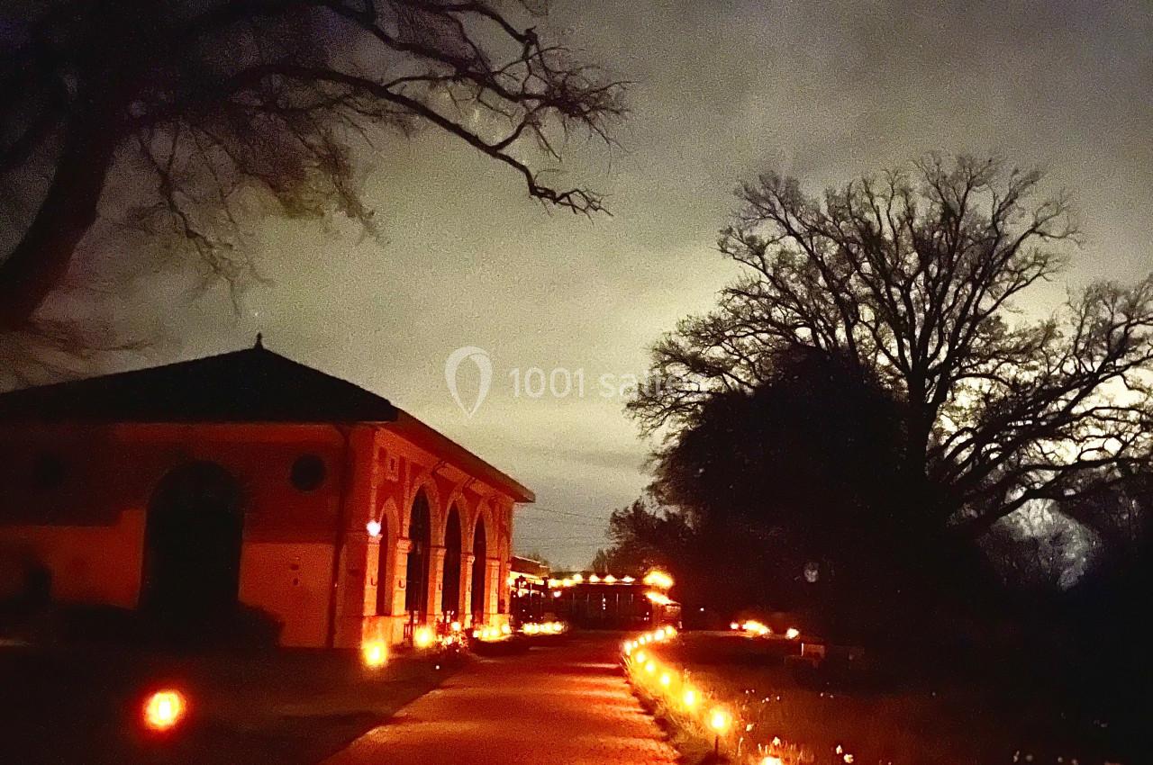 Allée pavée éclairée par des lanternes, bordée d'un bâtiment en briques et d'arbres sous un ciel nocturne.