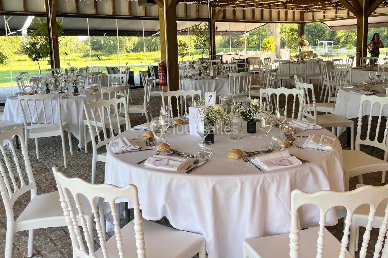 Salle de réception décorée avec des tables rondes dressées, nappes blanches, chaises élégantes et vue sur un jardin…