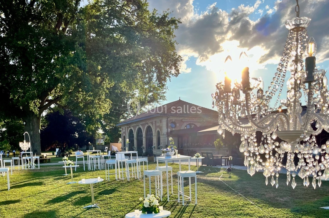 Jardin aménagé pour un événement avec tables hautes, chandeliers et bâtiment en arrière-plan sous un ciel ensoleillé.