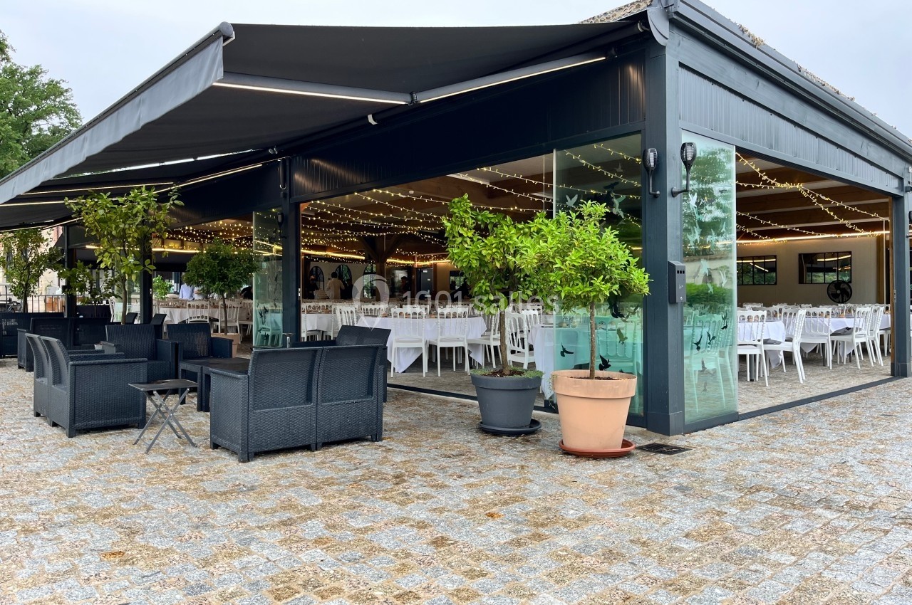 Terrasse couverte avec mobilier de jardin, entourée de plantes en pot, donnant sur une salle aménagée pour un événement.