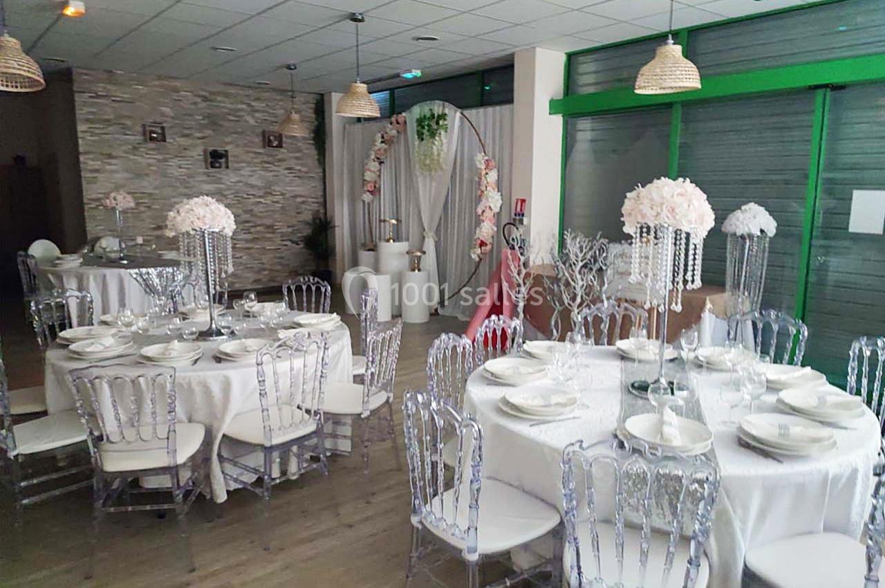 Salle de réception décorée avec des tables rondes, nappes blanches, chaises transparentes et arrangements floraux.