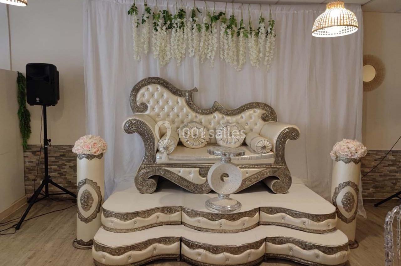 Canapé décoratif argenté et blanc sur une estrade, entouré de fleurs et d'une draperie blanche avec guirlandes suspendues.