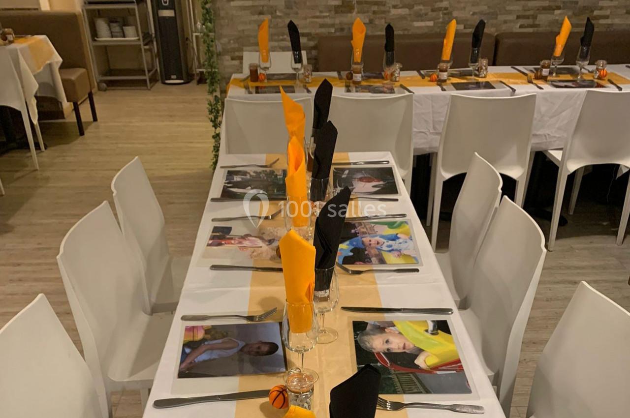 Tables dressées avec nappes blanches, serviettes orange et noires pliées, et sets de table illustrés dans une salle décorée.