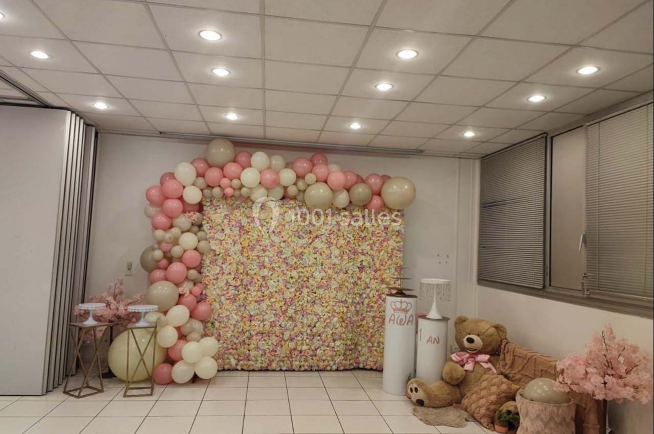Décoration festive avec mur de fleurs, ballons roses et blancs, peluches et éléments décoratifs dans une salle éclairée.