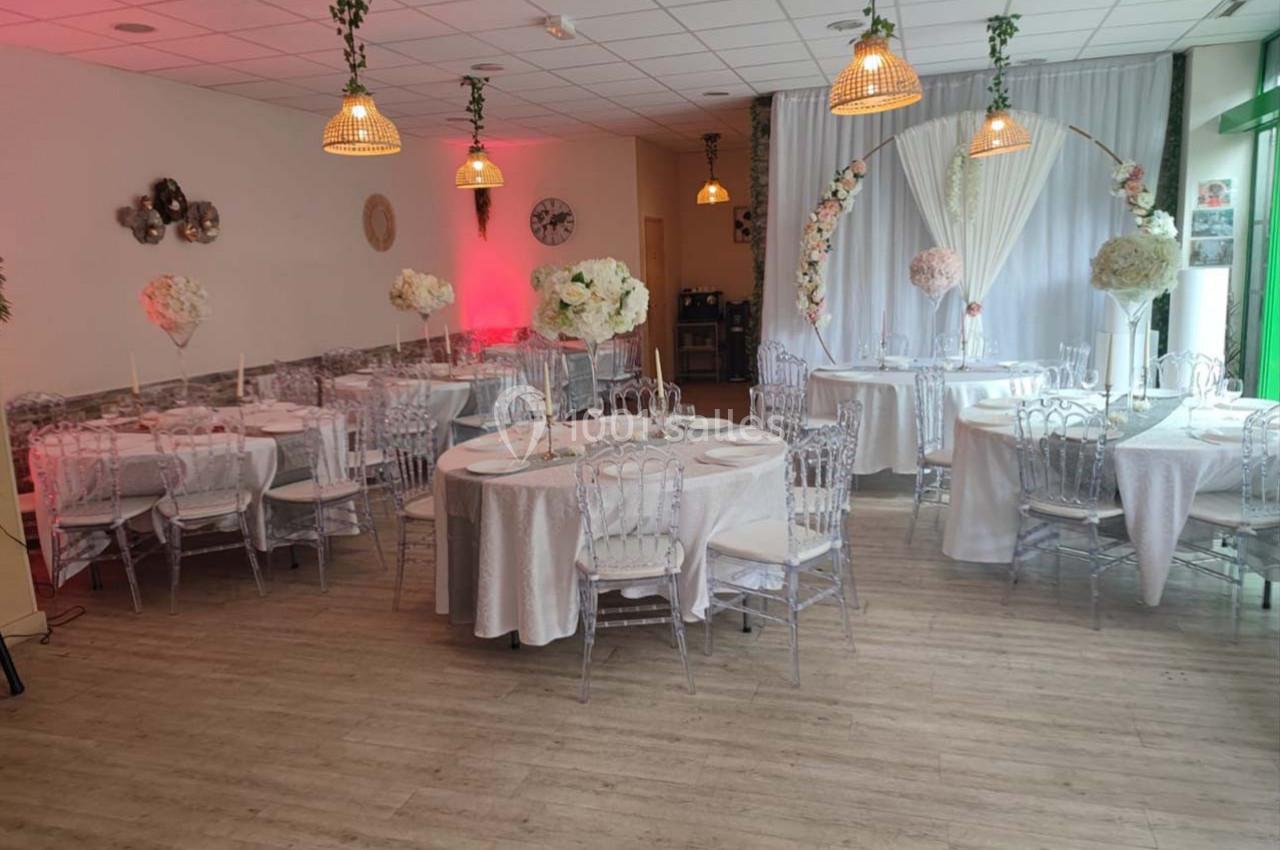 Salle décorée pour un événement avec tables rondes, nappes blanches, chaises transparentes et arche florale.