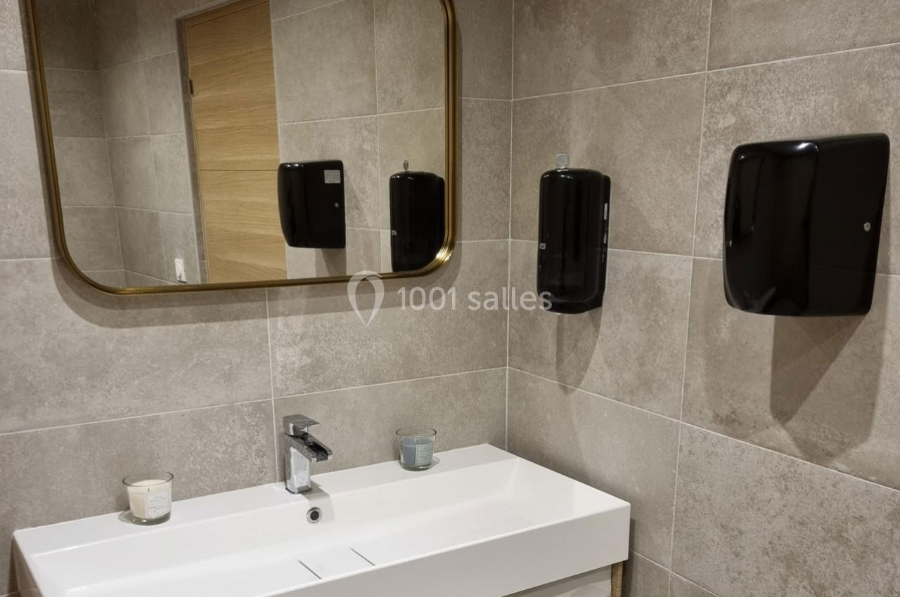 Lavabo blanc avec miroir rectangulaire doré, distributeurs noirs et murs en carrelage beige.