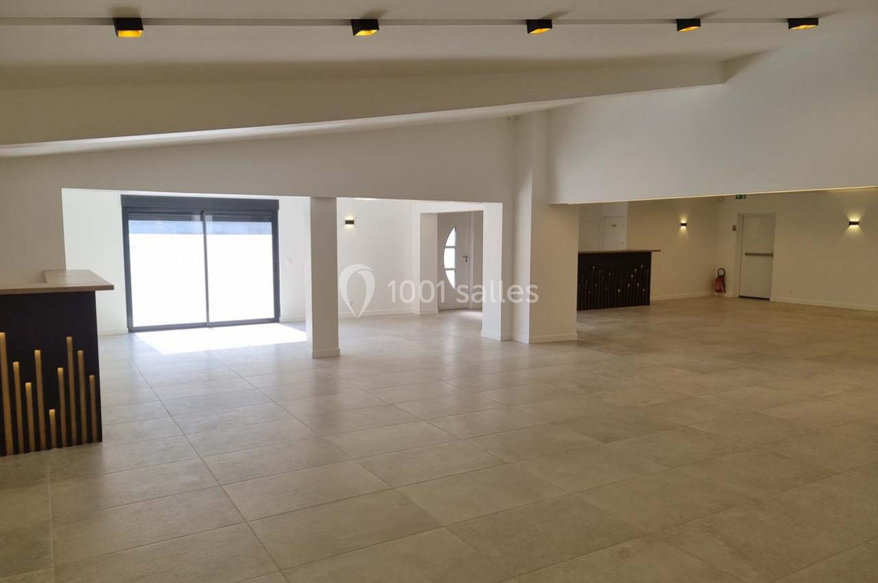 Salle spacieuse et lumineuse avec carrelage clair, bar à gauche, grandes baies vitrées et éclairage au plafond.