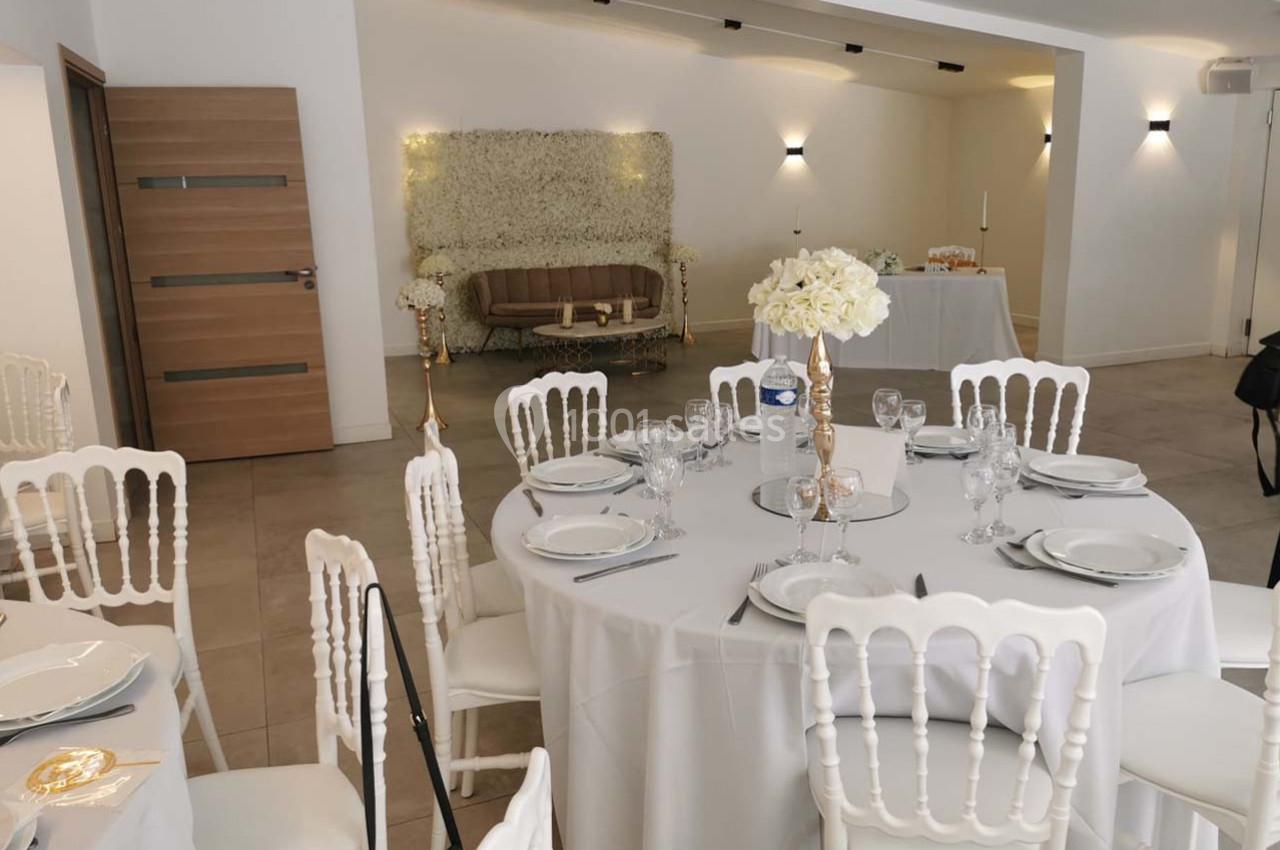 Salle de réception décorée avec des tables rondes dressées, des chaises blanches et un mur floral en arrière-plan.