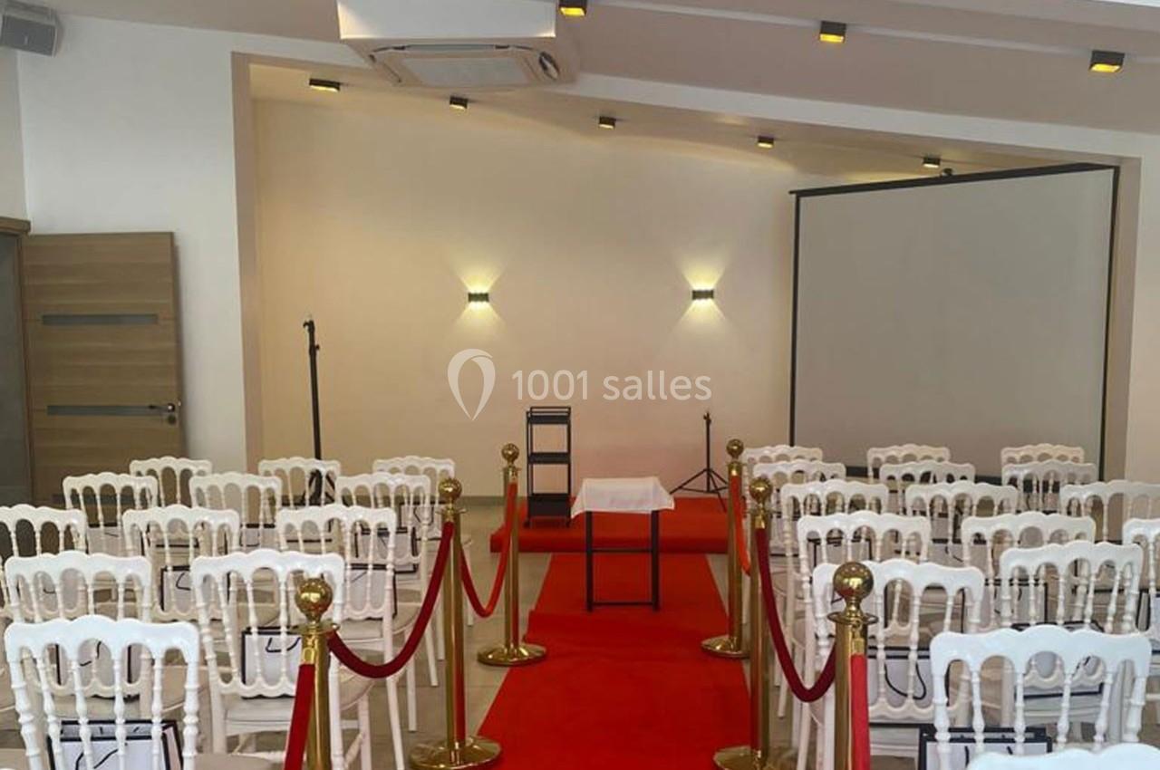 Salle aménagée avec des chaises blanches, tapis rouge, pupitre et écran de projection, prête pour un événement.