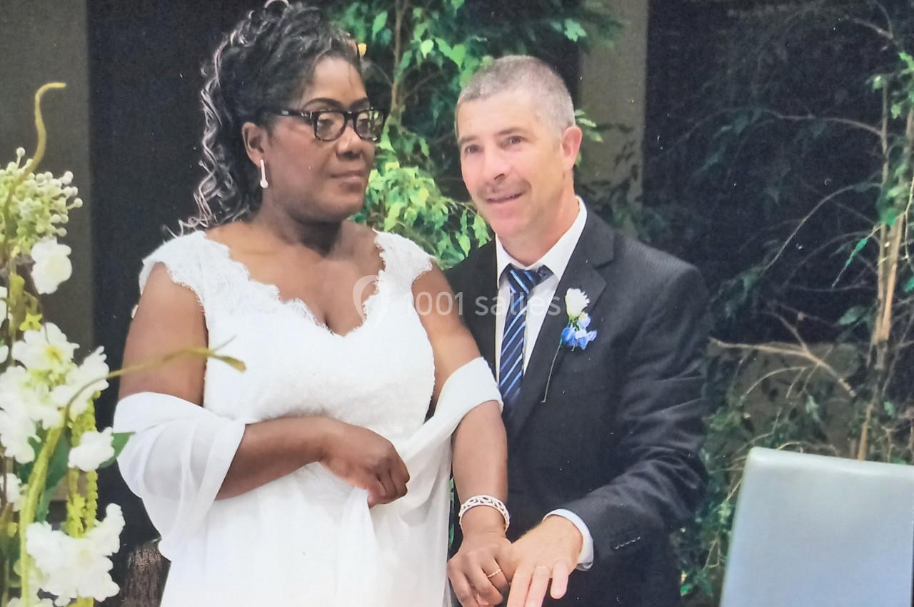 Un couple en tenue de mariage pose devant un décor végétal, la mariée portant une robe blanche.