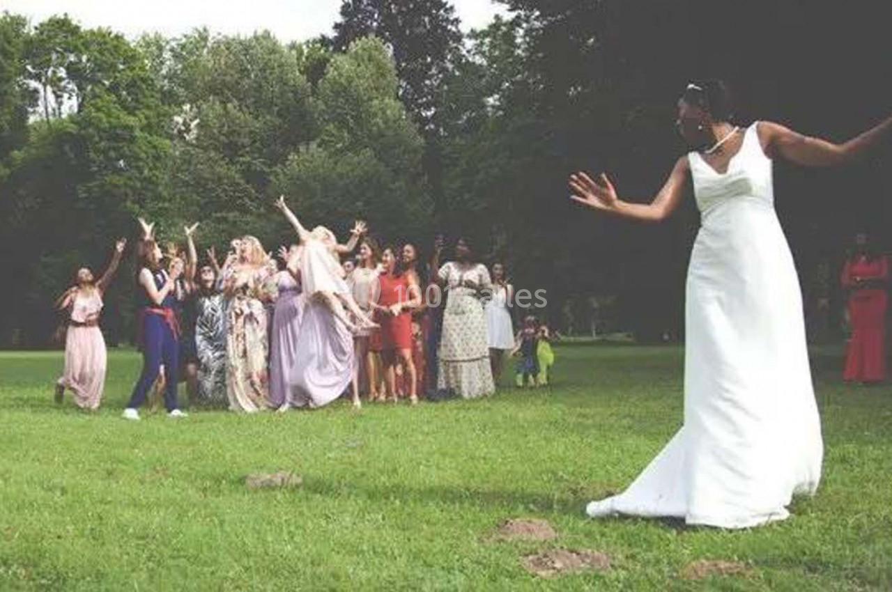 Une mariée lance son bouquet à un groupe de femmes rassemblées sur une pelouse dans un parc verdoyant.