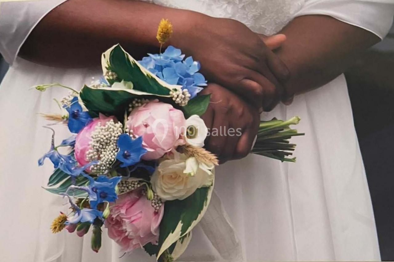 Une personne en robe blanche tient un bouquet de fleurs colorées composé de roses, pivoines et fleurs bleues.