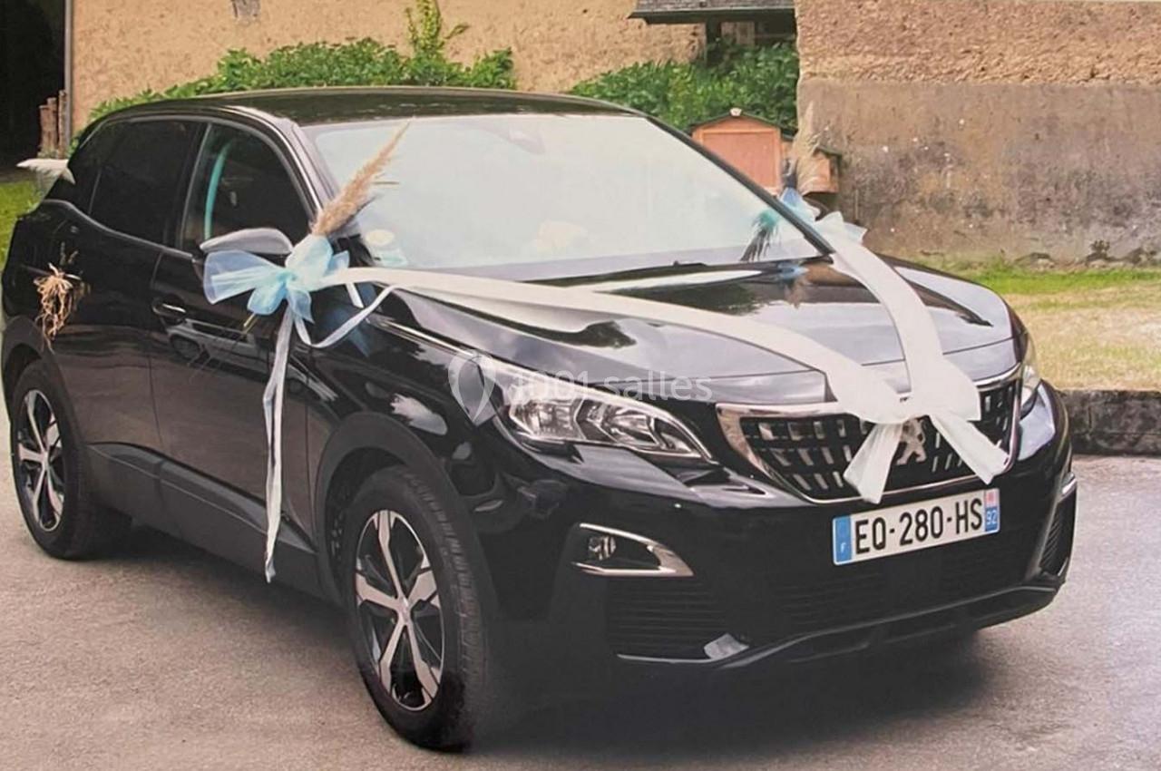 Voiture noire décorée de rubans blancs et bleus pour une cérémonie, stationnée devant un bâtiment en pierre.