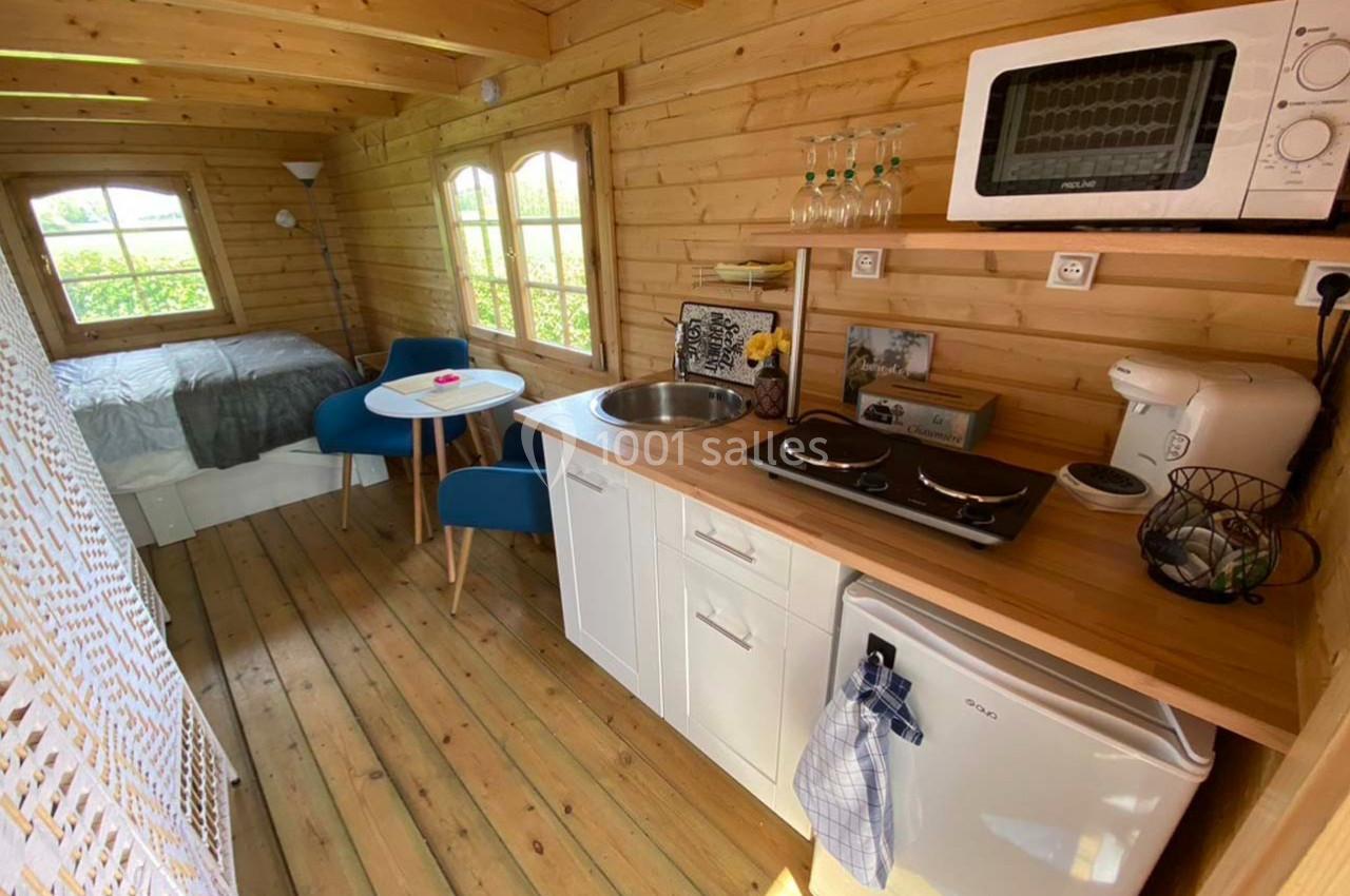 Intérieur d'une petite maison en bois avec kitchenette équipée, table, chaises et lit visible en arrière-plan.