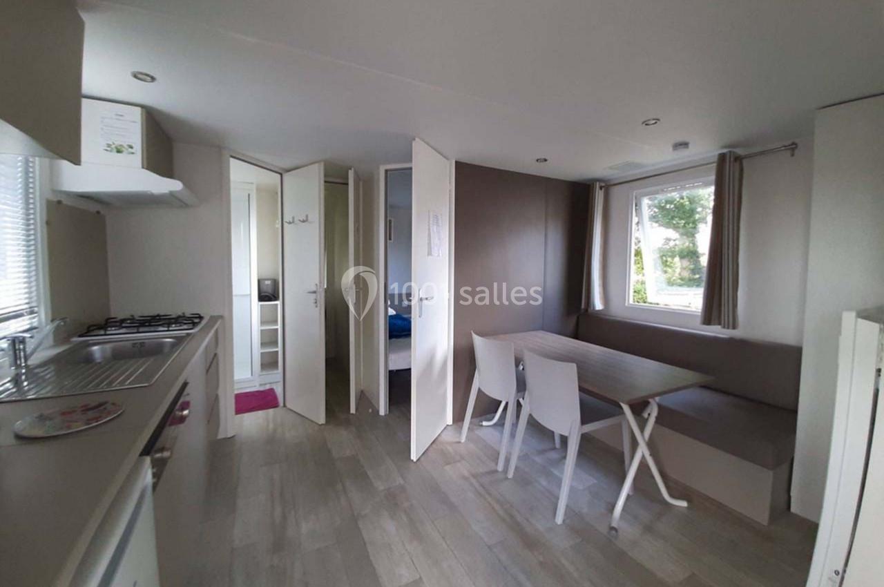 Intérieur d'un mobil-home avec coin cuisine, table à manger, banquette et vue sur une chambre à travers une porte ouverte.