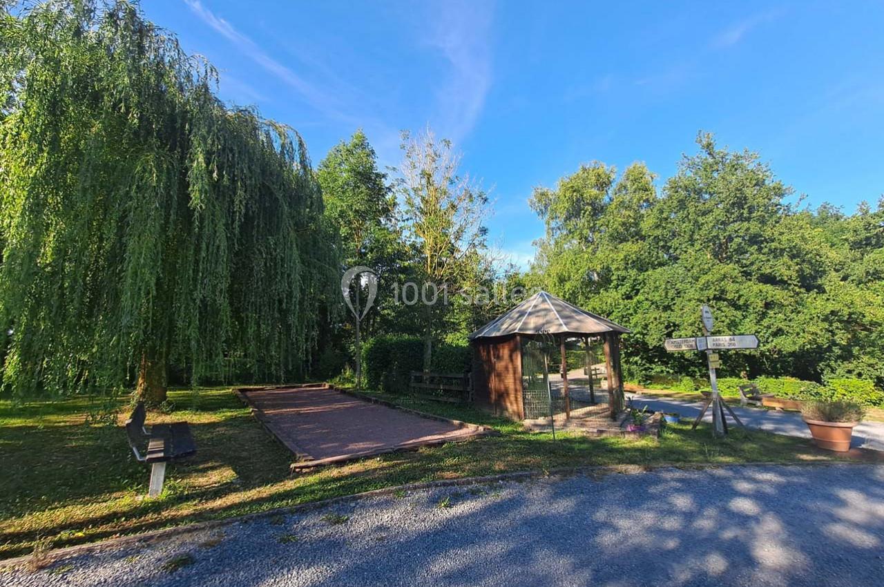 Kiosque en bois près d'un terrain de pétanque, entouré d'arbres et d'un chemin ensoleillé.