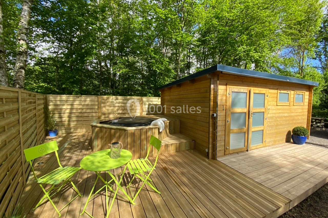 Terrasse en bois avec un jacuzzi, une table et des chaises vertes, entourée de verdure et d'une cabane en bois.