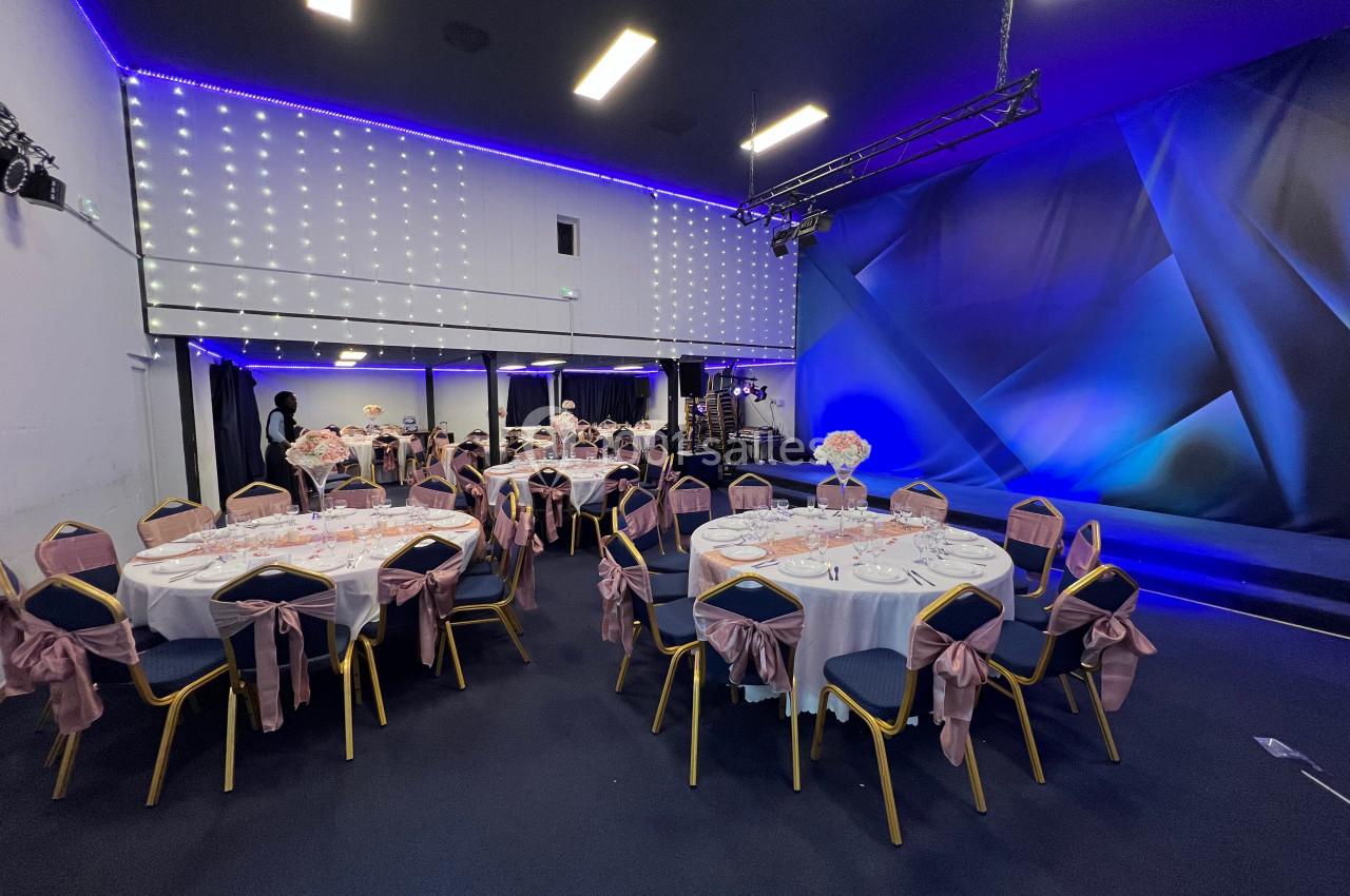 Salle de réception décorée avec des tables rondes dressées, nappes blanches et chaises ornées de nœuds roses.