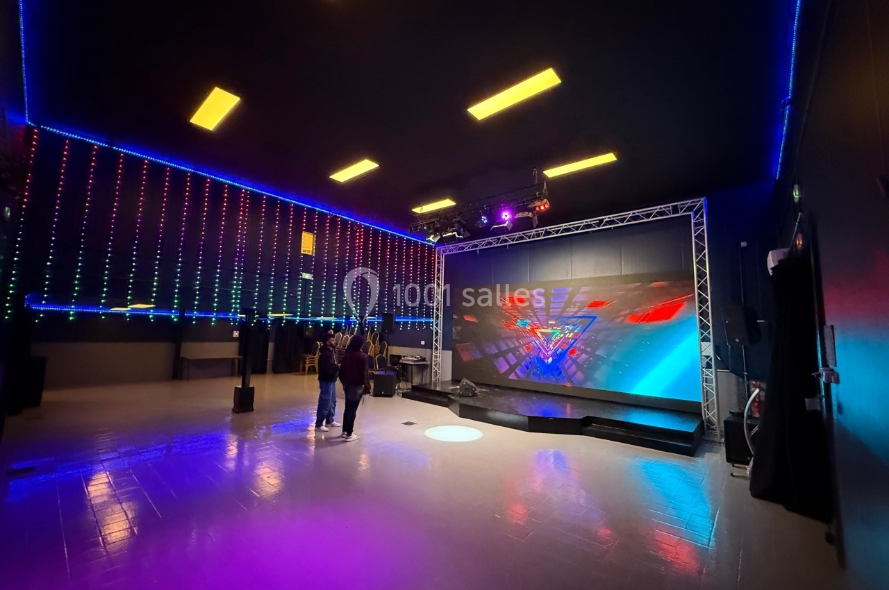 Salle avec un écran géant affichant des graphismes colorés, éclairages LED au plafond et quelques personnes présentes.