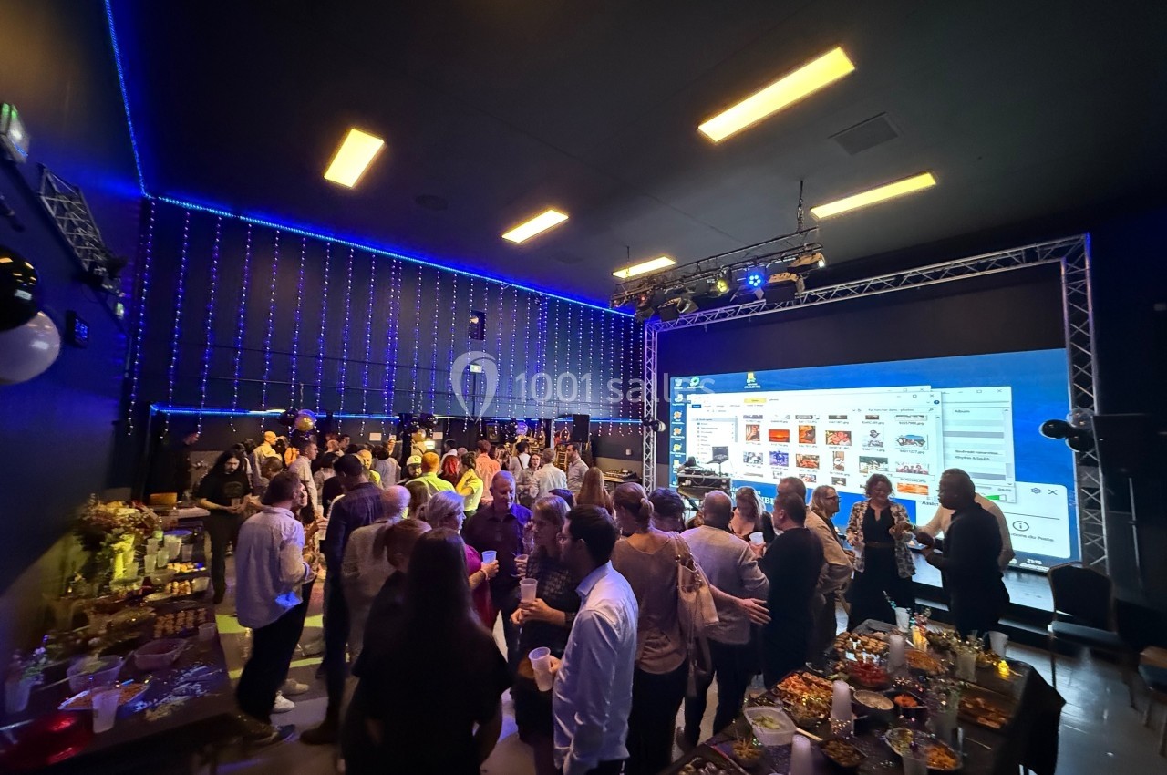 Groupe de personnes rassemblées dans une salle sombre avec éclairage bleu, buffet et grand écran en arrière-plan.