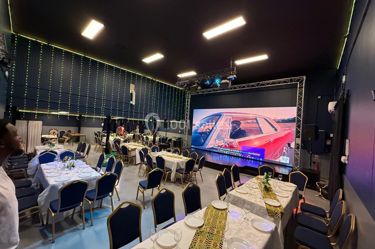 Salle décorée avec des tables dressées, chaises alignées et un grand écran affichant une voiture rouge.