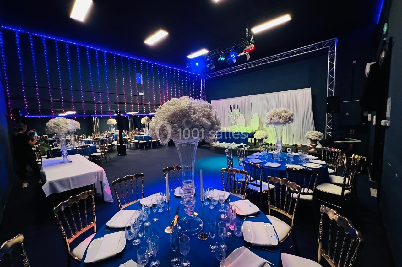 Salle de réception décorée avec des tables rondes, nappes bleues, chaises dorées et centres de table floraux blancs.
